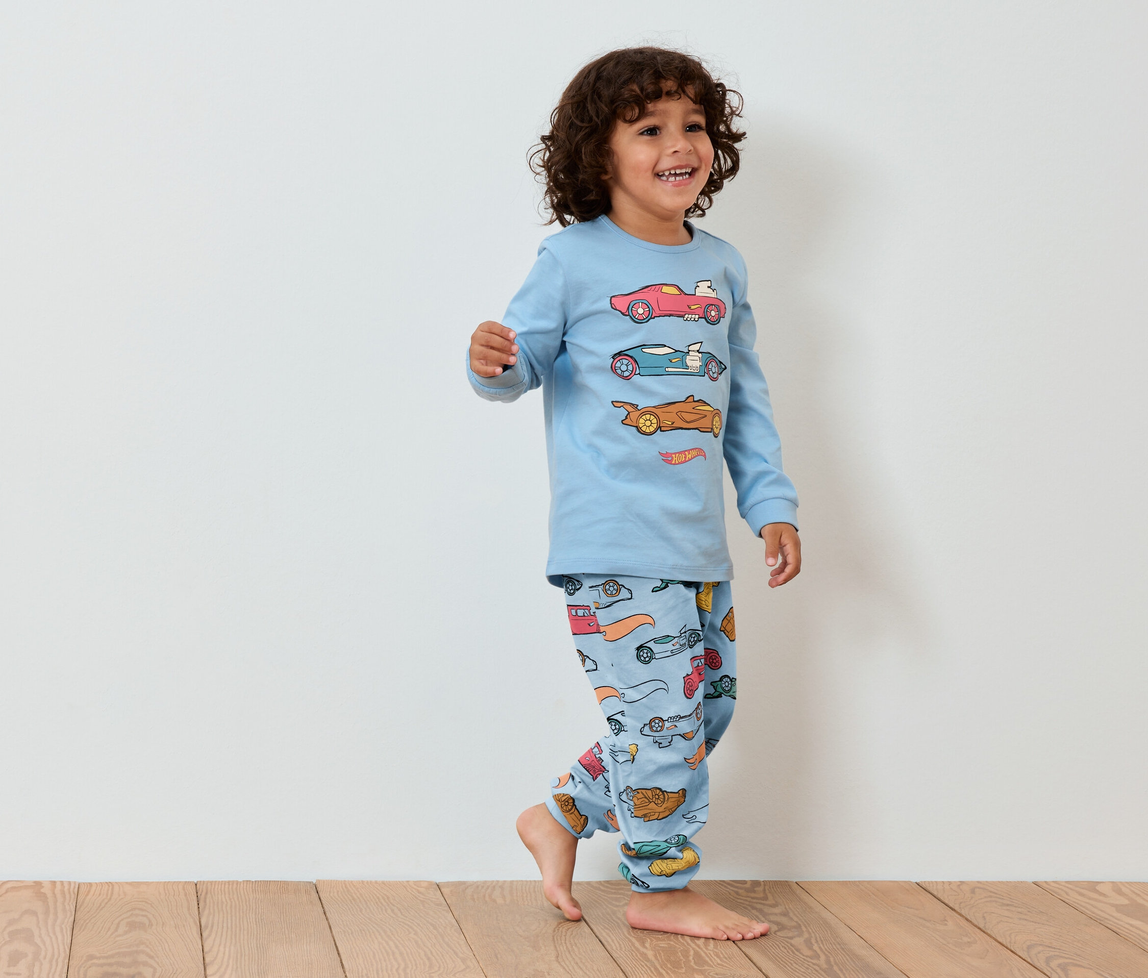 Lächelnder kleiner Junge in blauem Auto-Pyjama geht auf Holzboden.