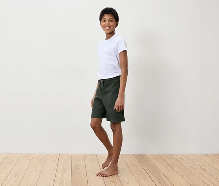 Ein Junge in einem weißen T-Shirt und grünen Shorts steht auf einem Holzboden vor einer weißen Wand.