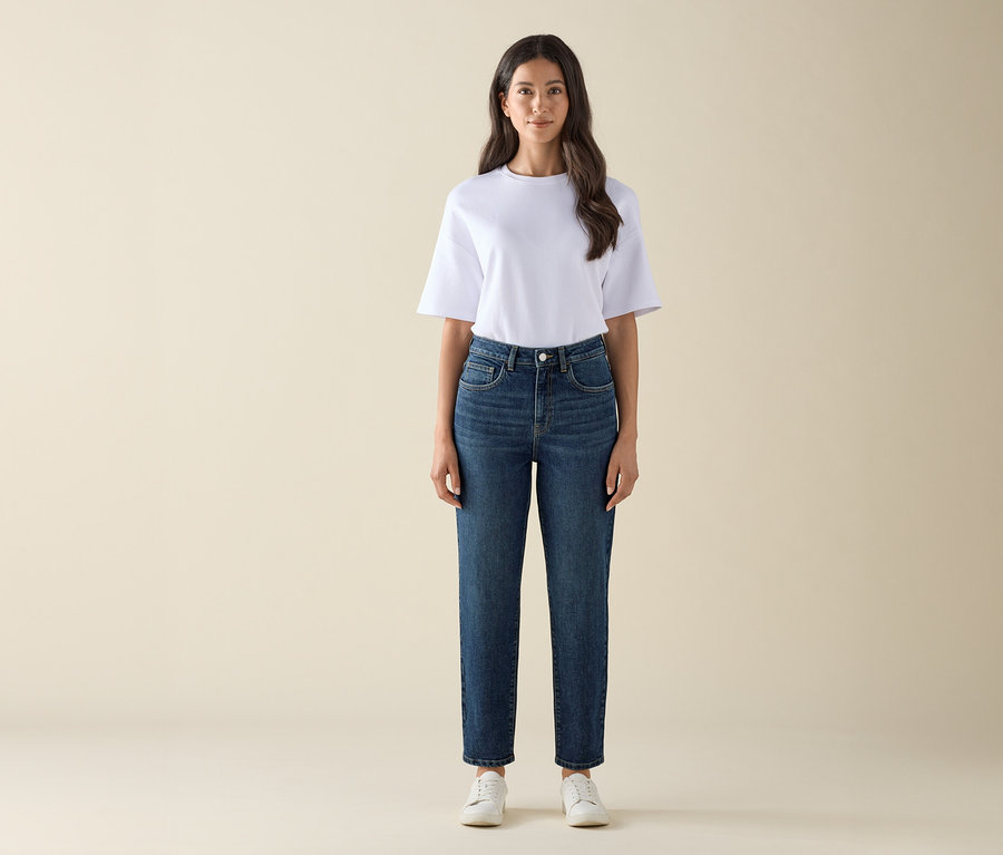 Frau steht und trägt eine Mom Jeans – Fit »Nyla« in Dunkelblau, ein weißes T-Shirt und weiße Turnschuhe.