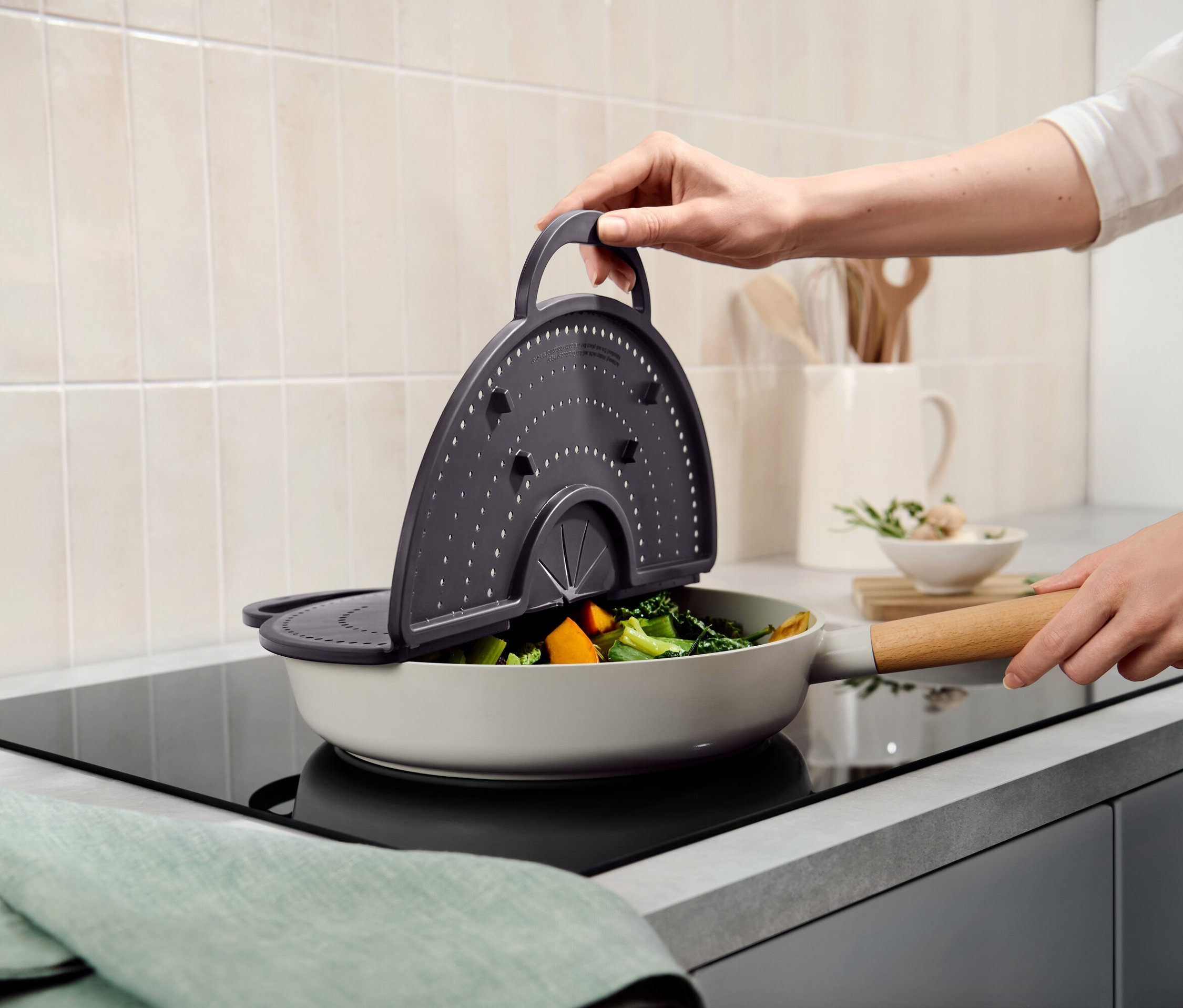 Eine Hand hält ein graues Sieb über eine Pfanne mit Gemüse.