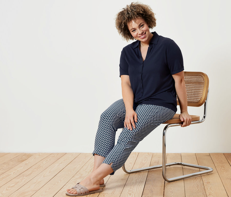 Eine Frau sitzt auf einem Stuhl und trägt eine dunkelblaue Blusenshirt mit Knopfleiste und eine bedruckte 7/8-Stretchhose.