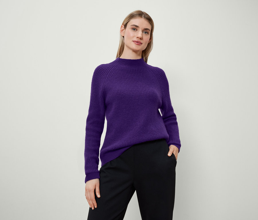 Frau posiert in lila Pullover und schwarzer Hose.