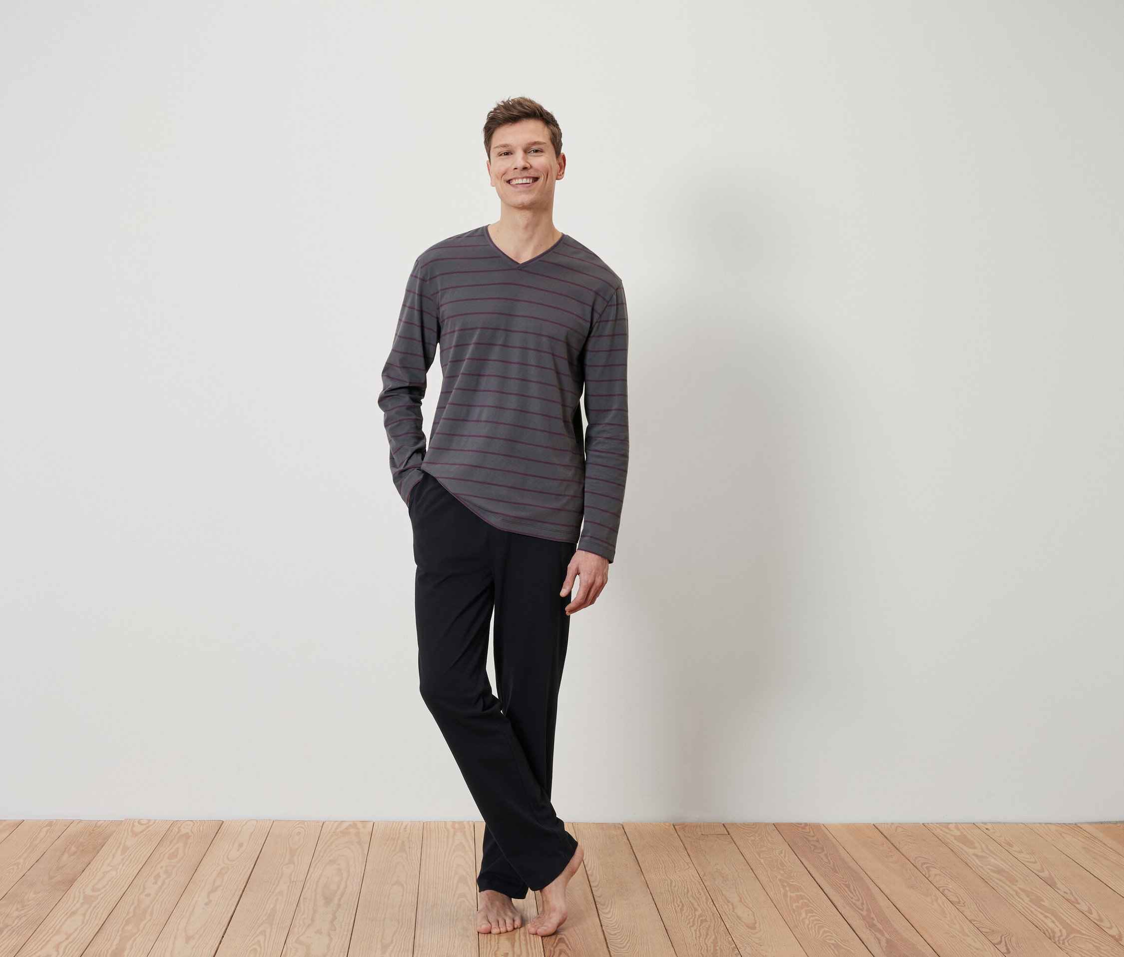 Mann steht barfuß auf Holzboden, bekleidet mit einem grau gestreiften Langarmshirt und einer schwarzen Hose.