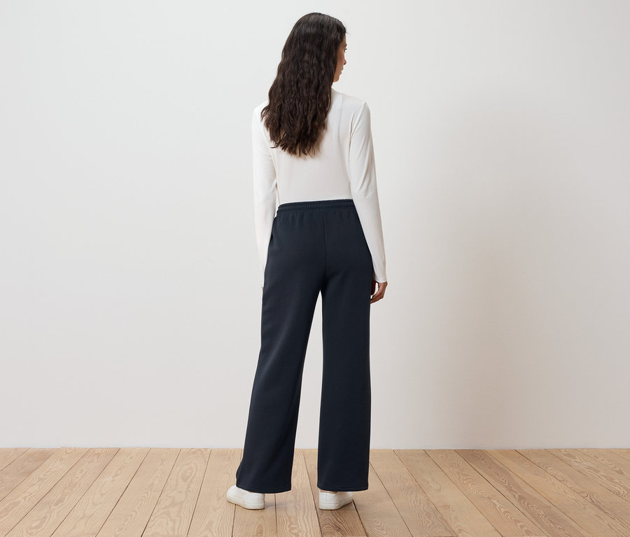 Frau mit langen, gewellten Haaren steht mit dem Rücken zur Kamera und trägt ein weißes Langarmshirt und eine dunkelblaue, weit geschnittene Hose.