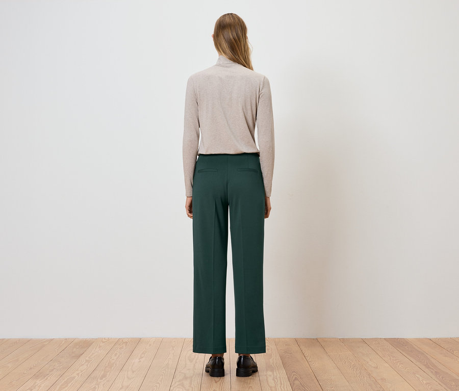 Frau steht mit dem Rücken zur Kamera, trägt einen beigen Rollkragenpullover und grüne Hosen.