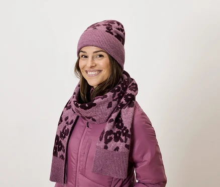Lächelnde Frau trägt eine lila Jacke, einen Schal mit Muster und eine passende Mütze.
