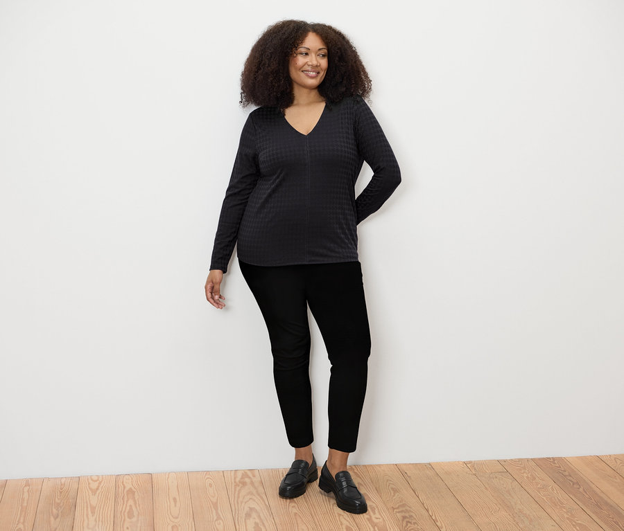 Eine Frau steht und trägt ein schwarzes, strukturiertes Langarmshirt, eine schwarze Hose und schwarze Schuhe.