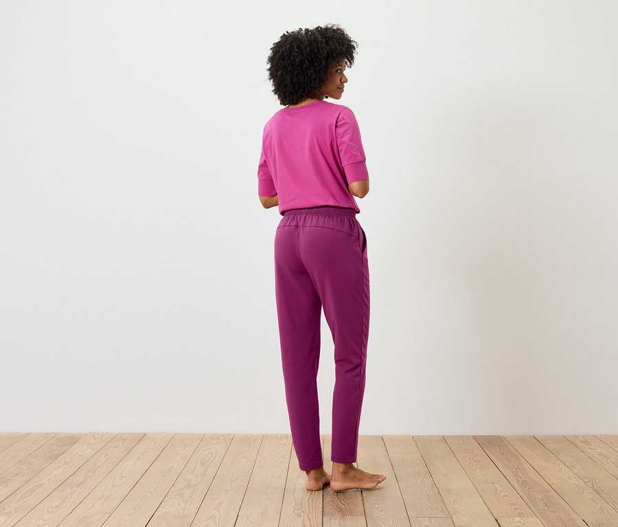 Frau mit Afro steht mit dem Rücken zum Betrachter und trägt ein pinkes T-Shirt und eine violette Hose.