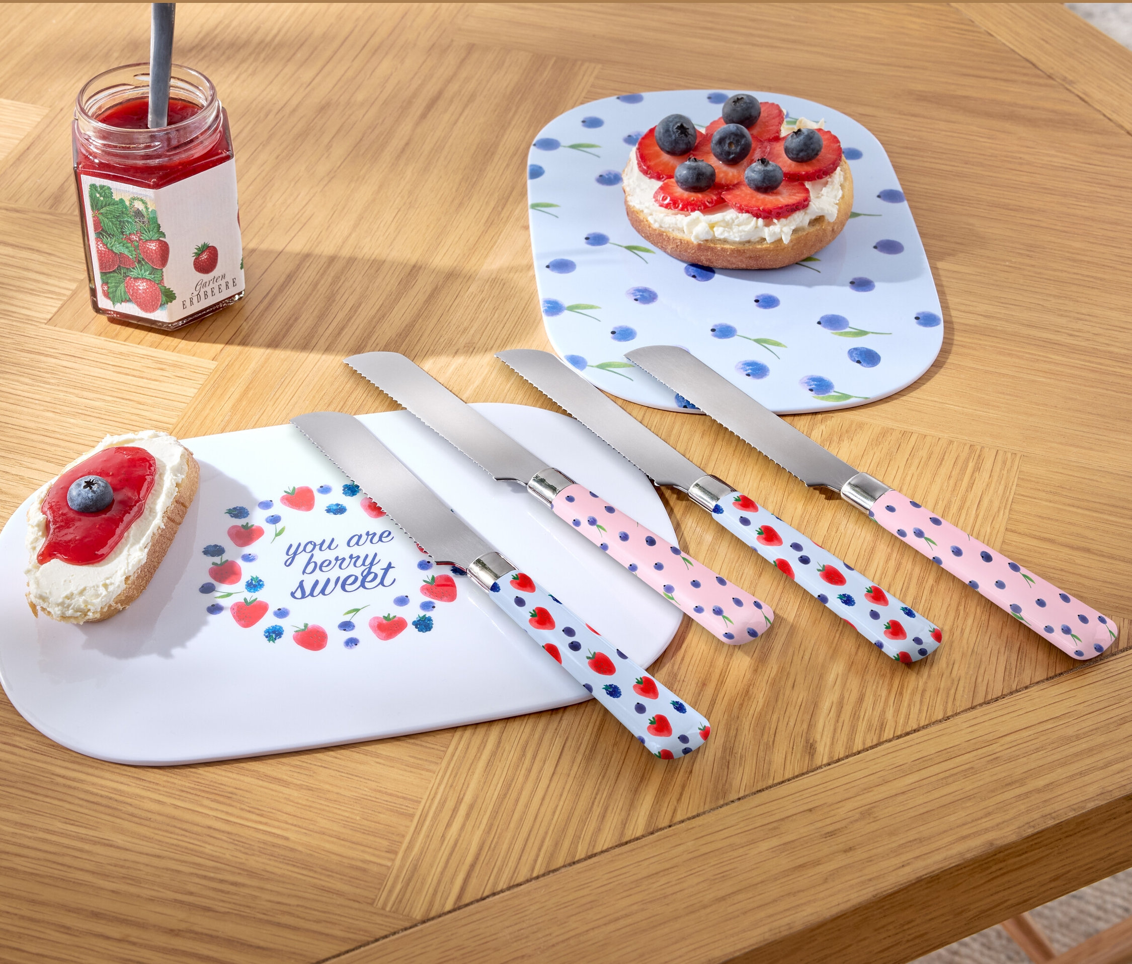 Vier Frühstücksmesser liegen auf einem Holztisch neben zwei Tellern und einem Glas Marmelade.