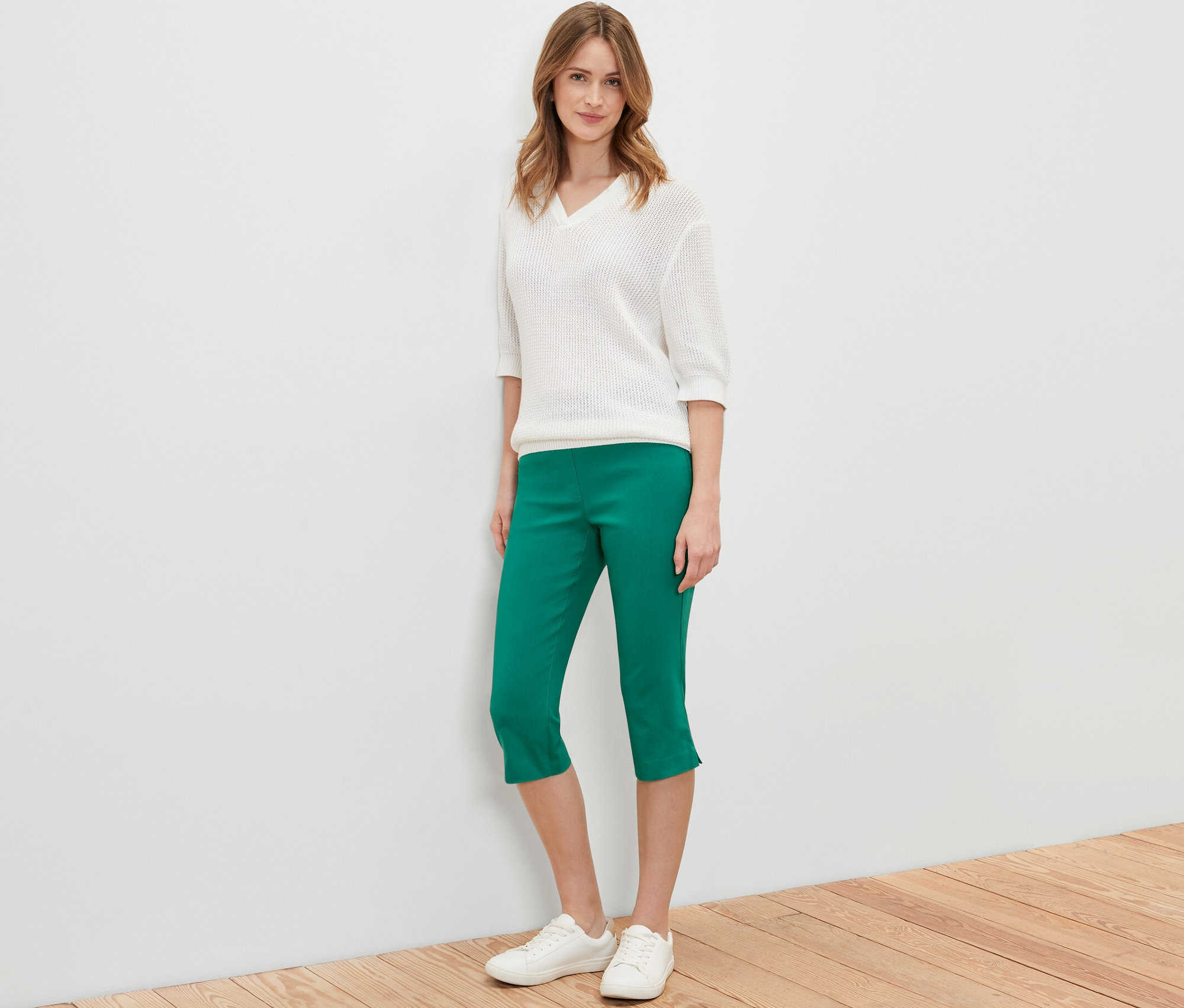 Frau posiert in grüner 3/4-Bengalin-Hose, weißem Pullover und weißen Schuhen.