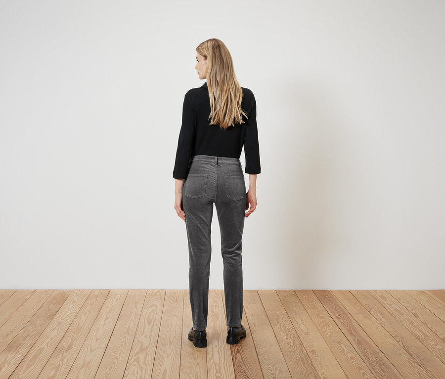 Frau steht mit dem Rücken, trägt ein schwarzes Hemd und graue Cordhosen.