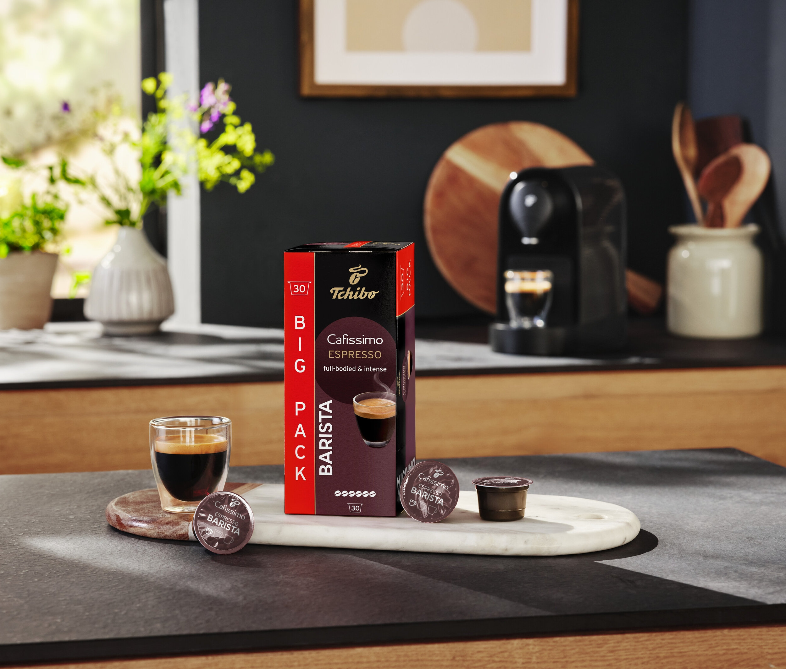 Ein Glas Espresso steht neben einer Packung Tchibo Cafissimo Barista Kapseln auf einem Küchentisch.