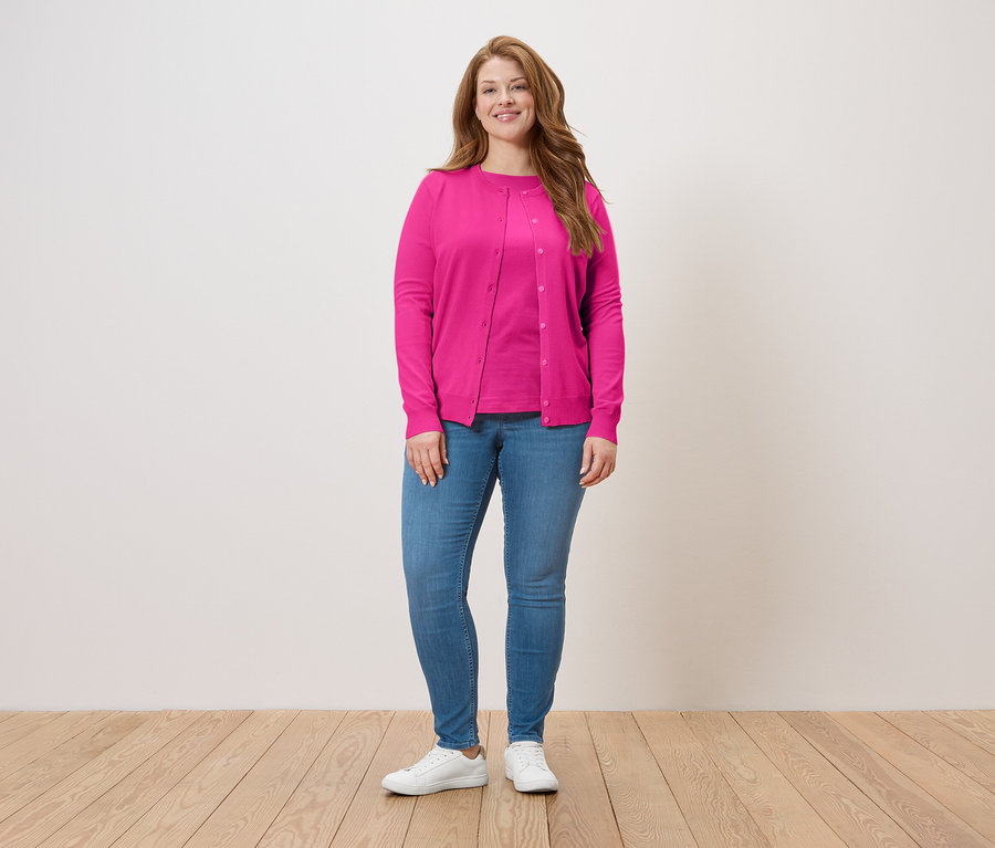 Eine Frau steht vor einer Wand und trägt eine pinke Strickjacke, ein pinkes T-Shirt, Jeans und weiße Turnschuhe.