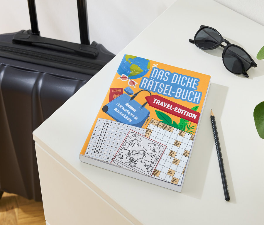 Reisekoffer steht neben Rätselbuch, Sonnenbrille und Bleistift auf einem Tisch.