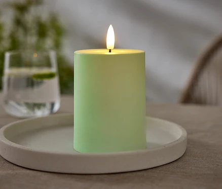 Eine grüne Kerze steht auf einem weißen Teller. Im Hintergrund ist ein Glas Wasser mit Limette zu sehen.