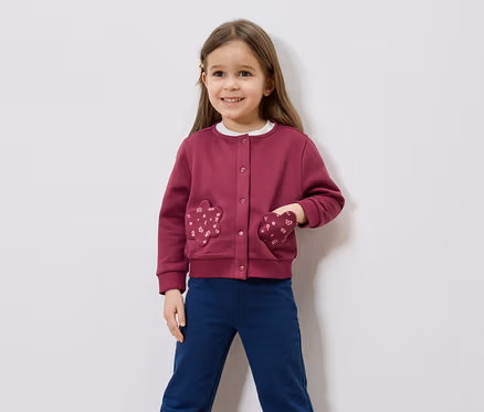 Lächelndes Mädchen mit braunen Haaren, trägt eine weinrote Strickjacke mit blumenförmigen Taschen und blaue Hosen vor einem weißen Hintergrund.