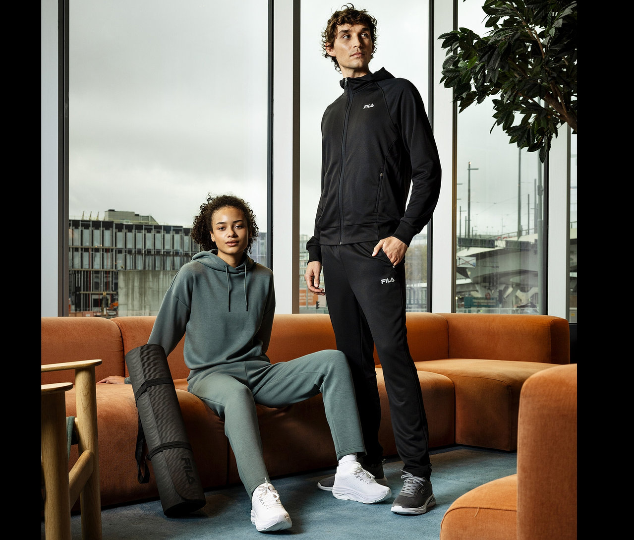 Eine junge Frau sitzt auf einem orangefarbenen Sofa und hält eine schwarze Yogamatte. Ein junger Mann in schwarzem Trainingsanzug steht daneben.