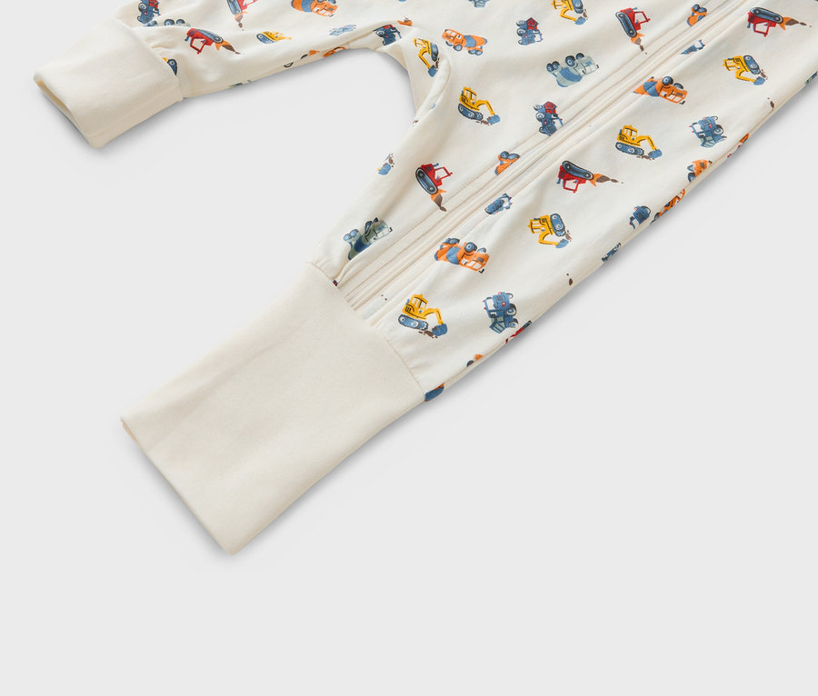 Detailansicht eines Baby-Schlafanzugs mit Reißverschluss und einem Muster aus verschiedenen Baufahrzeugen auf beigem Hintergrund.