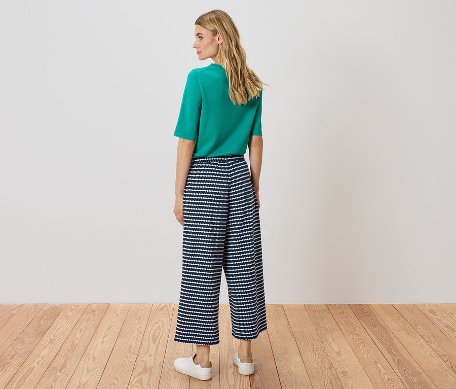 Eine Frau steht mit dem Rücken zur Kamera, sie trägt ein grünes Oberteil und eine blau-weiss gestreifte Culotte-Hose.