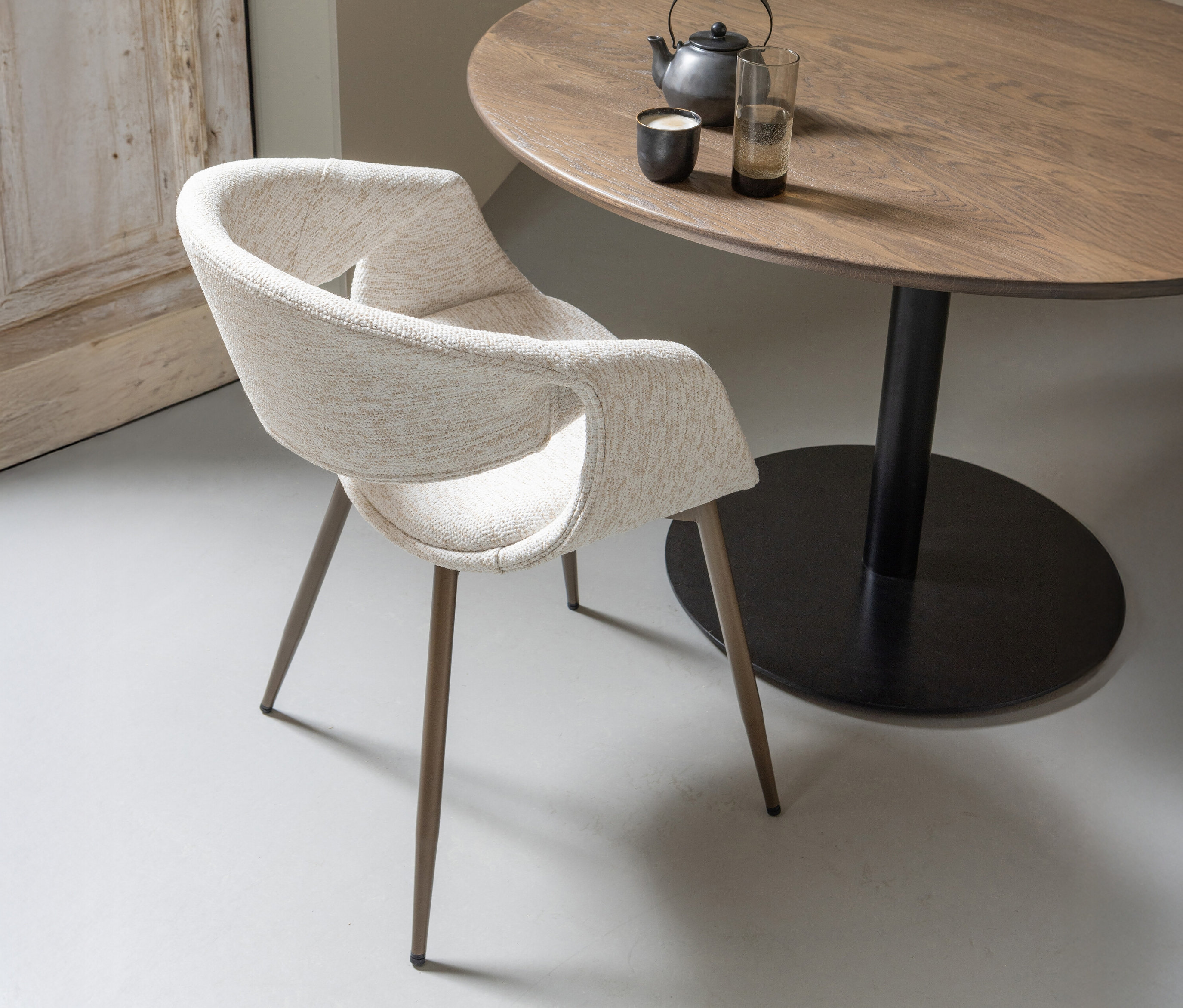 Ein kupfer-naturfarbener WOOOD Esszimmerstuhl »Yossi« mit Metallfuß aus einem 2er-Set steht neben einem runden Holztisch mit Teekanne und Tasse.