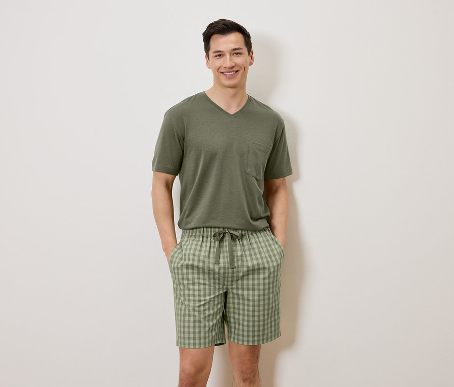Ein Mann steht mit den Händen in den Taschen vor einer weißen Wand, er trägt ein olivgrünes T-Shirt und karierte Shorts.
