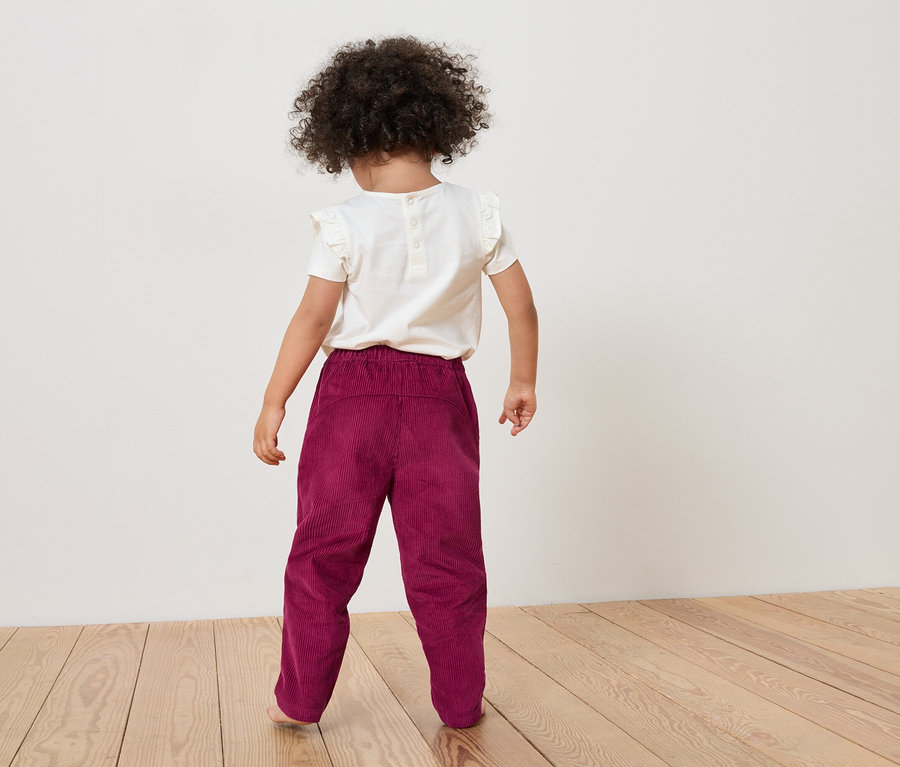 Rückansicht eines Kindes mit lockigem Haar, das ein weißes T-Shirt und eine weinrote Cordhose auf einem Holzboden trägt.