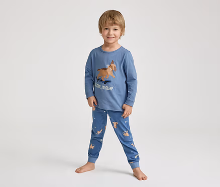 Ein lächelnder kleiner Junge steht barfuß in einem blauen Schlafanzug mit einem Hund auf einem Skateboard-Aufdruck und der Aufschrift "TOO COOL TO SLEEP".