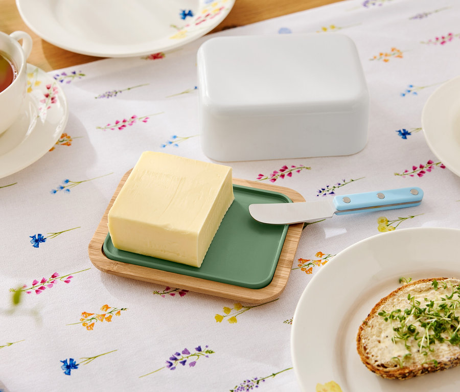 Auf einem blumigen Tisch liegt eine Butter auf einem hölzernen Teller mit grünem Einsatz, ein Buttermesser, eine Butterdose, eine Tasse Tee und ein Teller mit einem Butterbrot.