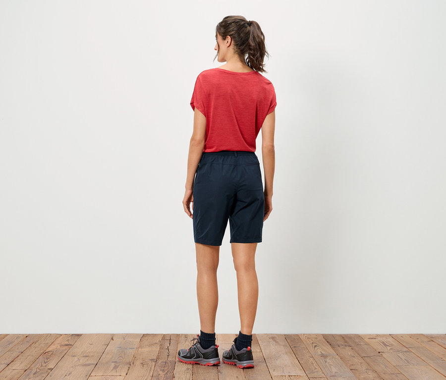 Rückansicht einer Frau mit rotem T-Shirt und blauen Shorts, die auf einem Holzboden steht.