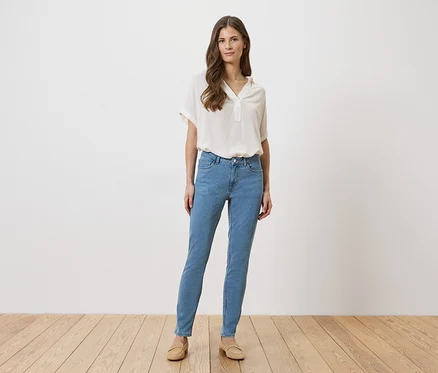 Frau in hellblauen Jeans, beiger Bluse und Schuhen steht vor einer weißen Wand auf einem Holzboden.