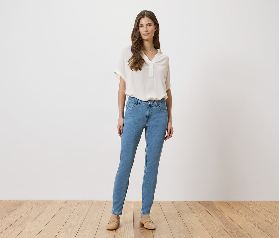 Frau in hellblauen Jeans, beiger Bluse und Schuhen steht vor einer weißen Wand auf einem Holzboden.