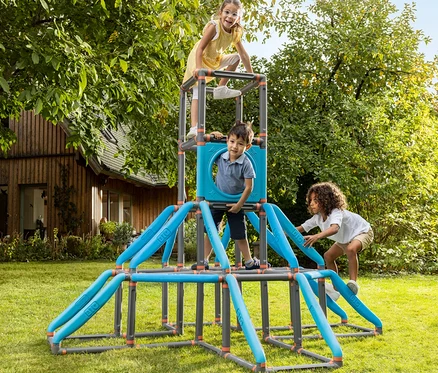 Drei Kinder spielen auf einem blauen und grauen Klettergerüst im Garten. Ein Mädchen steht oben, ein Junge klettert, ein anderes Mädchen klettert hoch.