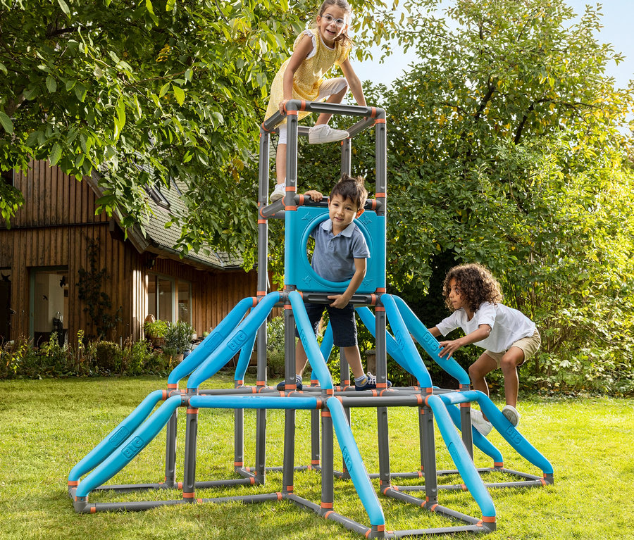 Drei Kinder spielen auf einem blauen und grauen Klettergerüst im Garten. Ein Mädchen steht oben, ein Junge klettert, ein anderes Mädchen klettert hoch.
