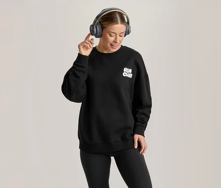 Eine Frau trägt ein schwarzes Sweatshirt mit „RUN CLUB“-Aufschrift, schwarze Leggings und graue Kopfhörer, lächelt und berührt ihren rechten Kopfhörer.