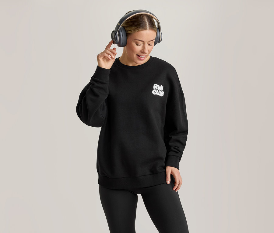 Eine Frau trägt ein schwarzes Sweatshirt mit „RUN CLUB“-Aufschrift, schwarze Leggings und graue Kopfhörer, lächelt und berührt ihren rechten Kopfhörer.