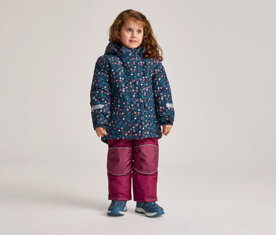 Ein kleines Mädchen mit lockigem Haar steht in einer blauen, geblümten Jacke und einer weinroten Hose.