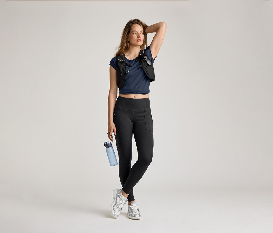 Frau in Outdoor-Leggings, blauem Shirt und Laufweste posiert mit Wasserflasche und Hand am Kopf.