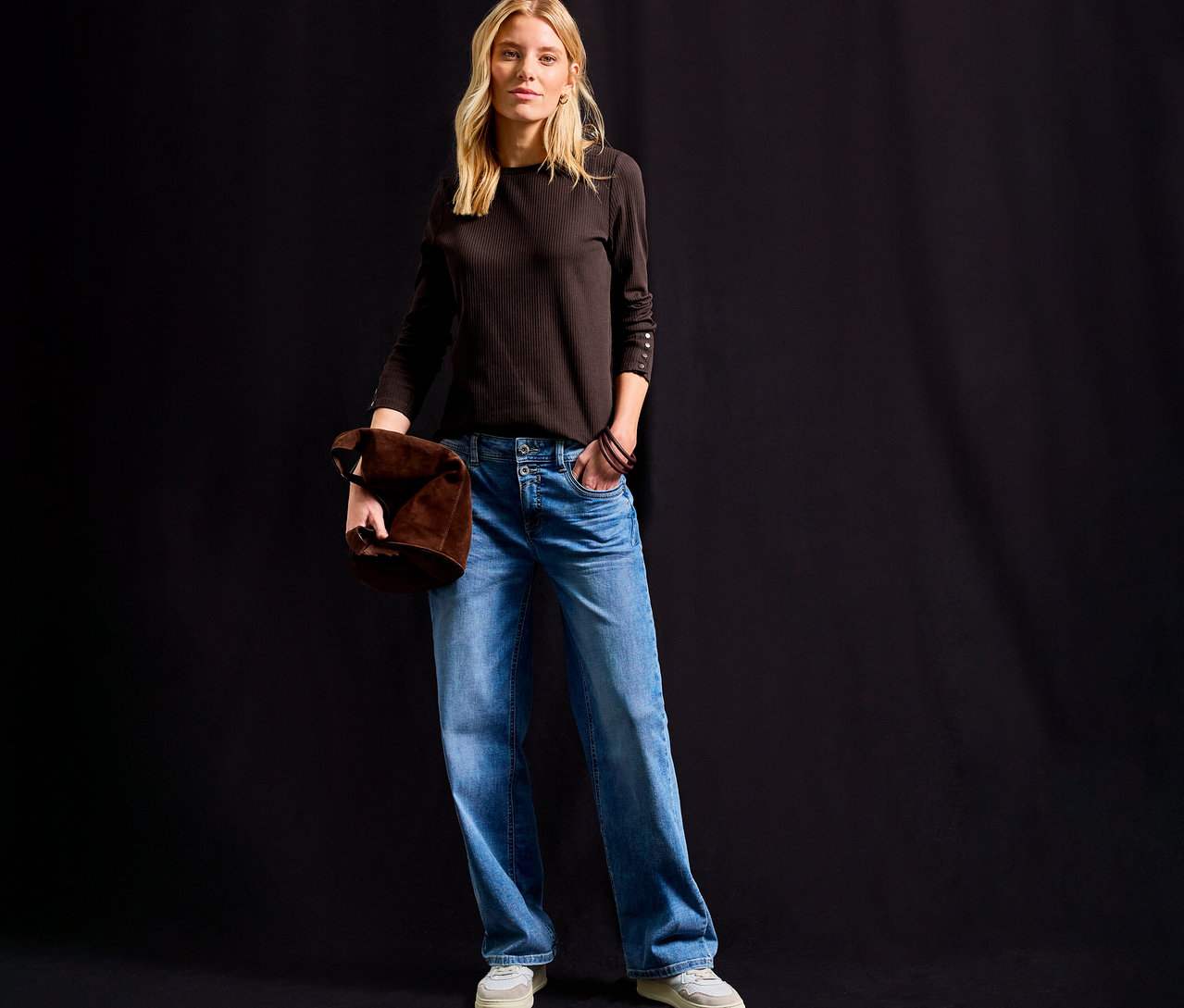 Frau steht und hält eine braune Tasche, trägt ein Street One Rippshirt in Braun, Jeans und helle Schuhe.