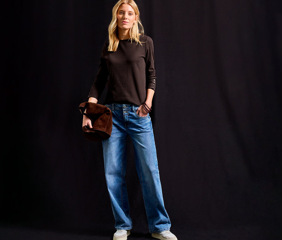 Frau steht und hält eine braune Tasche, trägt ein Street One Rippshirt in Braun, Jeans und helle Schuhe.