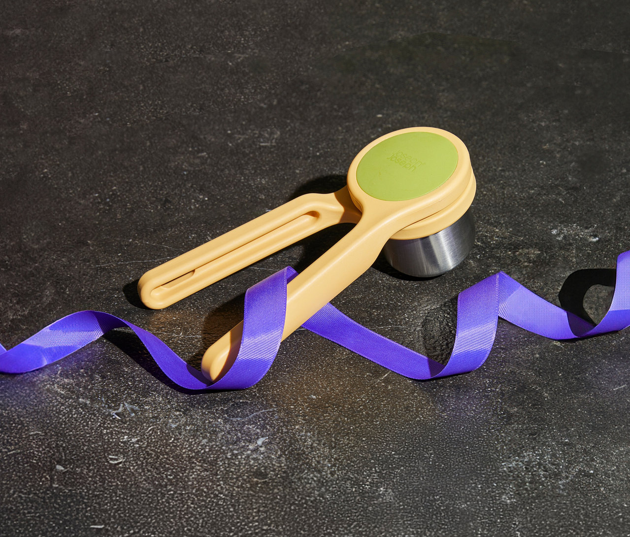 Eierschneider mit hellgelbem Griff und hellgrünem Einsatz liegt auf dunklem Untergrund, umwickelt von blauem Band.