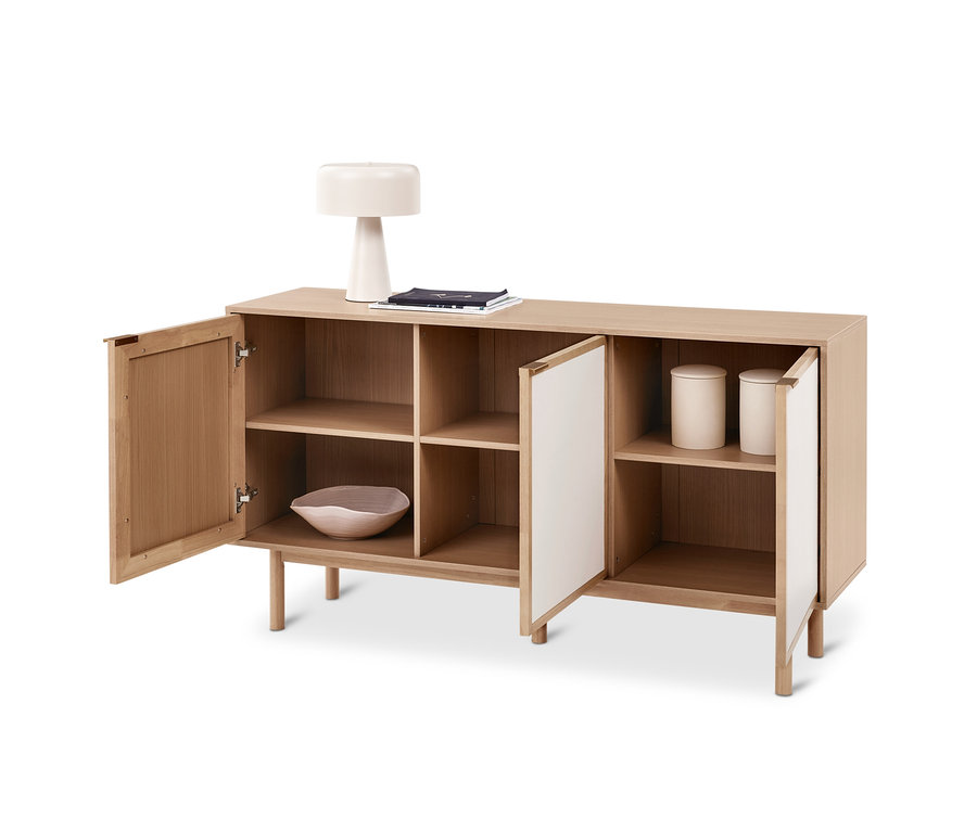 Helles Holz-Sideboard mit vier Fächern, die äußeren Türen sind offen. Obenauf eine weiße Lampe und Bücher, innen eine Schale und zwei Behälter.