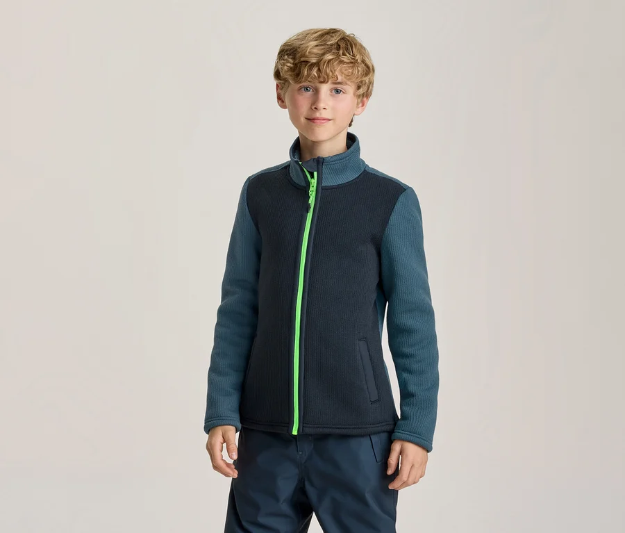 Junger Junge steht und blickt in die Kamera. Er trägt eine dunkelblaue Jacke mit türkisen Ärmeln und einem neongrünen Reißverschluss, dazu eine dunkelblaue Hose.