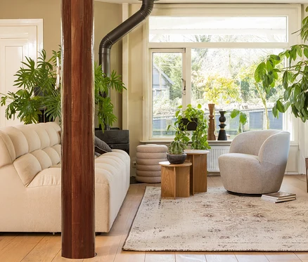 Ein gemütliches Wohnzimmer mit einem cremefarbenen, getufteten Sofa, einem hellgrauen Sessel und zwei Holztischen mit Pflanzen. Eine große Holzsäule steht im Vordergrund, und ein Holzofen ist hinter dem Sofa sichtbar. Gestapelte beigefarbene Poufs stehen am Fenster, das eine grüne Außenansicht bietet. Ein gemusterter Teppich bedeckt den Holzfußboden.