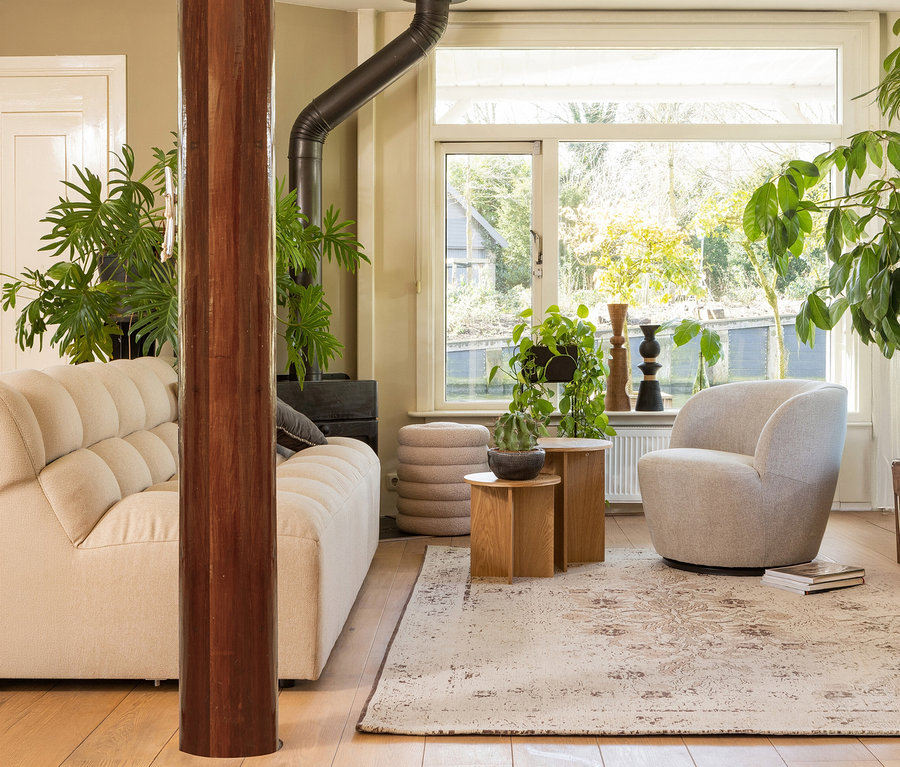 Ein gemütliches Wohnzimmer mit einem cremefarbenen, getufteten Sofa, einem hellgrauen Sessel und zwei Holztischen mit Pflanzen. Eine große Holzsäule steht im Vordergrund, und ein Holzofen ist hinter dem Sofa sichtbar. Gestapelte beigefarbene Poufs stehen am Fenster, das eine grüne Außenansicht bietet. Ein gemusterter Teppich bedeckt den Holzfußboden.