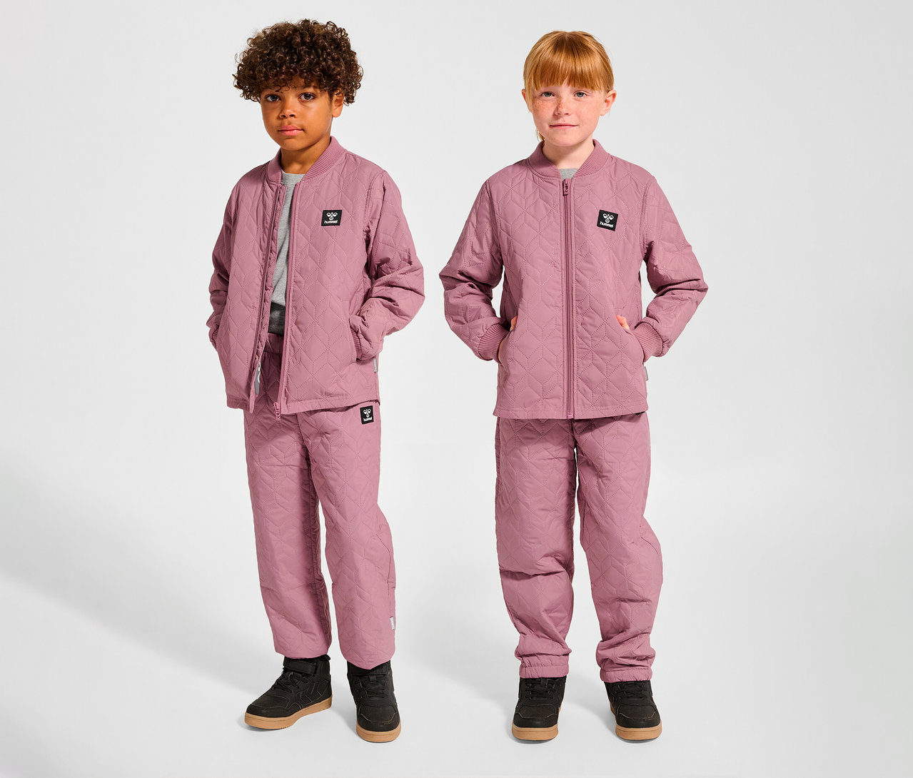 Zwei Kinder, ein Junge und ein Mädchen, stehen nebeneinander und tragen passende rosa hummel® HMLSOBI Thermo-Sets, bestehend aus gesteppten Jacken und Hosen.