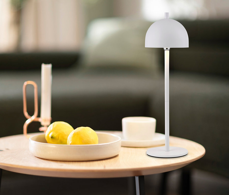 Weiße SCHÖNER WOHNEN-Kollektion Akku-Tischleuchte »Bellis« auf einem Holztisch neben einer Schale mit Zitronen und einer Kerze.