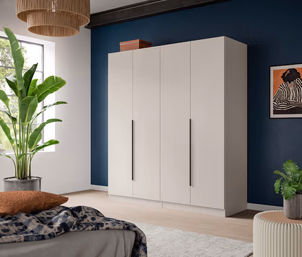 Ein modernes Zimmer mit einem großen, hellbeigen viertürigen Kleiderschrank an einer dunkelblauen Wand. Im Vordergrund befindet sich ein Bett mit einer gemusterten Decke und einem braunen Kissen. Eine hohe grüne Pflanze steht an einem Fenster, und eine kleinere Pflanze auf einem runden Beistelltisch. Ein gerahmtes Kunstwerk hängt an der blauen Wand.