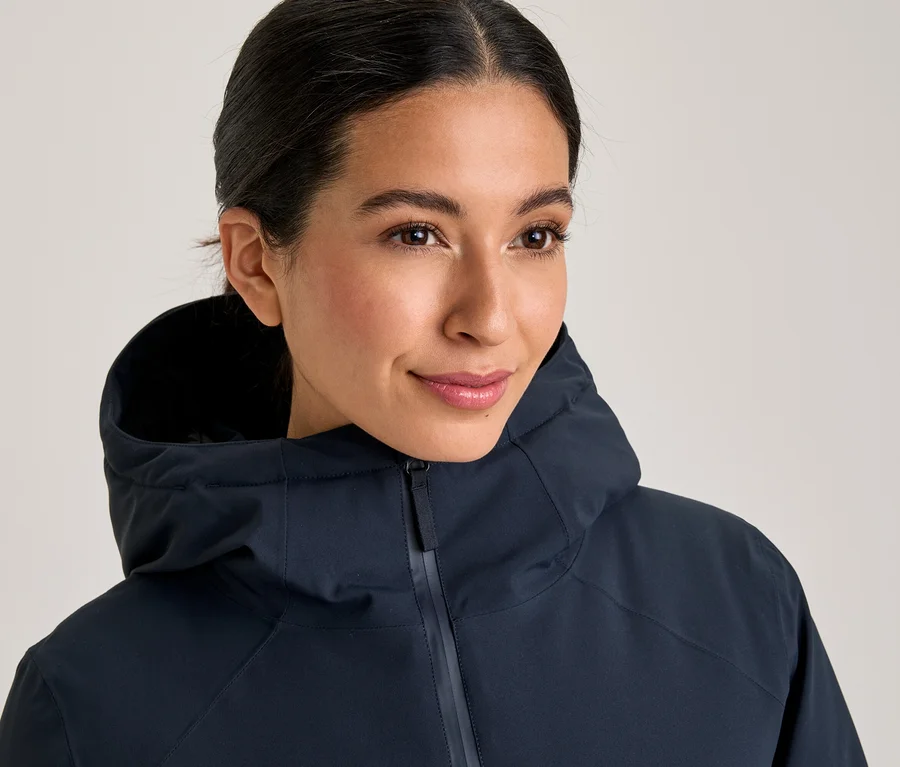 Eine lächelnde Frau mit zurückgebundenem dunklem Haar trägt eine dunkelblaue Kapuzenjacke, die bis zum Hals geschlossen ist.