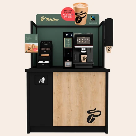 Tchibo Kaffeestation mit schwarzem und holzfarbenem Schrank, dunkelgrüner Rückwand und Kaffeemaschine, die in einen Becher füllt. Ein Schild bewirbt "Großer Kaffee, kleiner Preis" neben einem Menü und Zubehör.