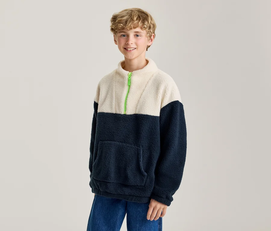 Lächelnder Junge trägt einen zweifarbigen Fleece-Pullover in Creme und Marineblau mit leuchtend grünem Reißverschluss und blauen Jeans.